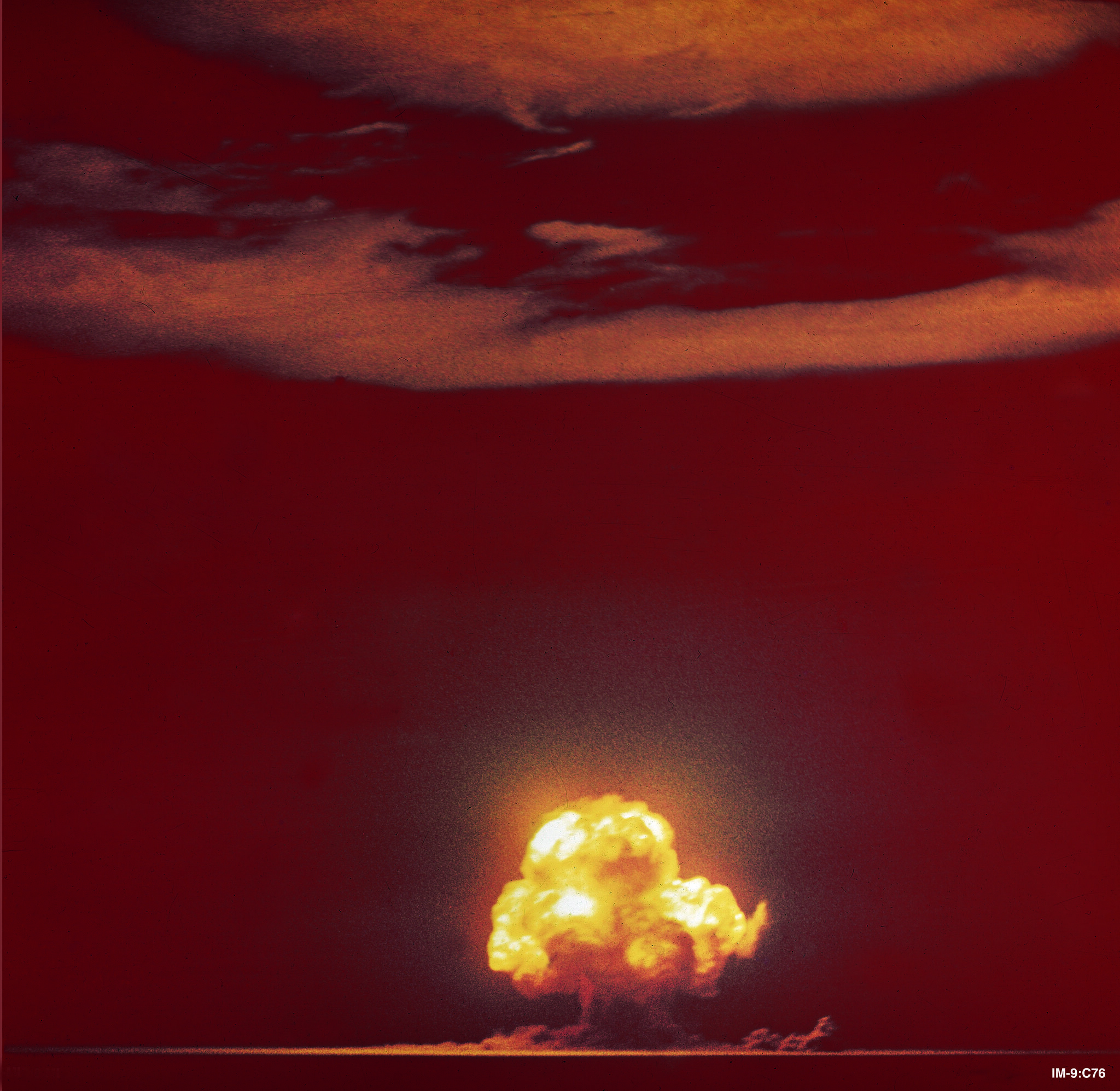 Reddish photo with a lit up mushroom cloud that glows yellow at the top. Nothing else is visible in the landscape. 