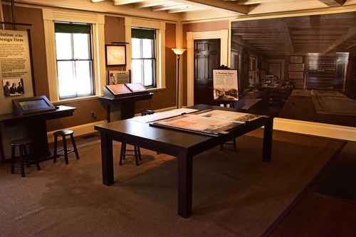 Large table in middle of room with exhibits on one side, and a black and white view of the original room. 