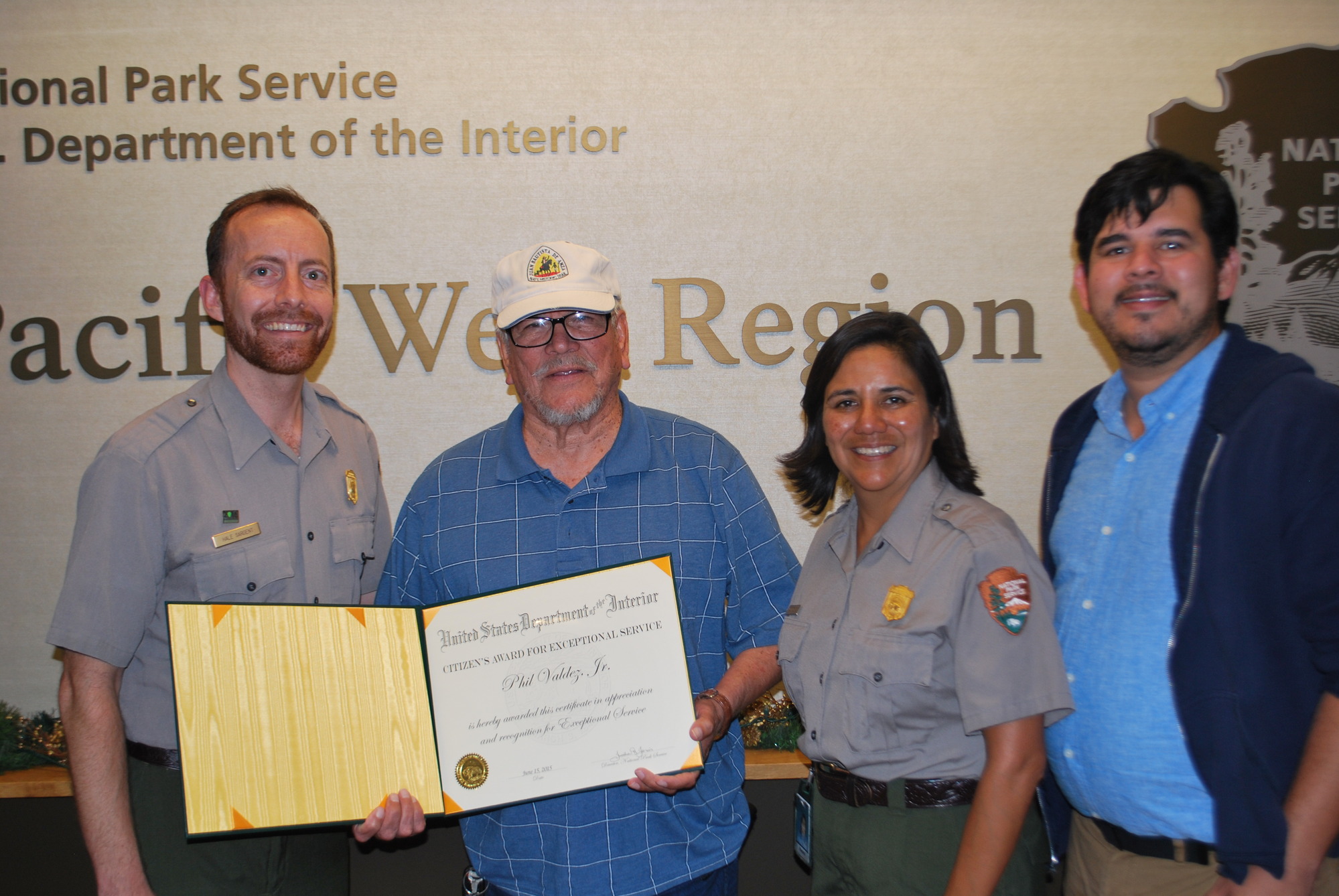 A man in glasses and an Anza Trail hat holds an award next to two NPS employees and a third person in a button-down shirt and sweatshirt in front of a wall that says National Park Service US Department of the Interior Pacific West Region
