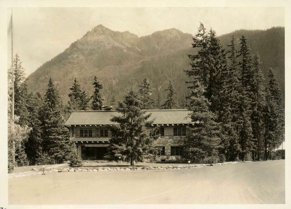 A two story wood and stone building framed by trees with a large dirt parking lot. A forested mountain peak rises in the background. 