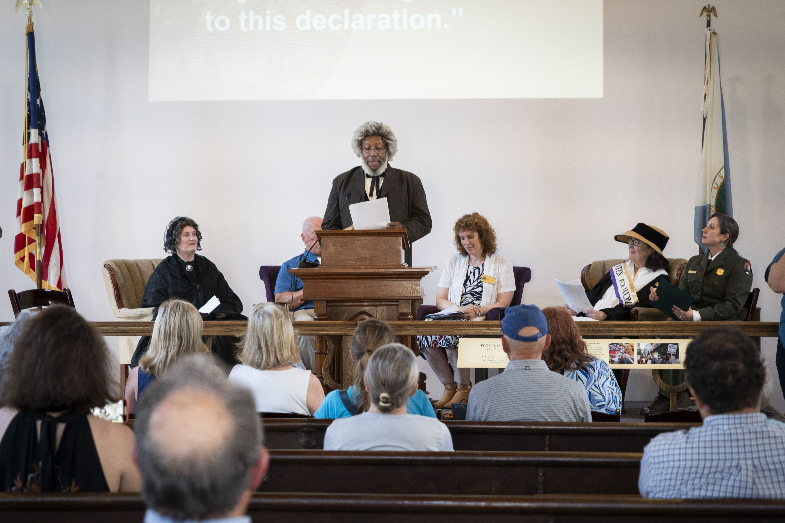 A man in a black suit and gray wig stands behind a podium and speaks