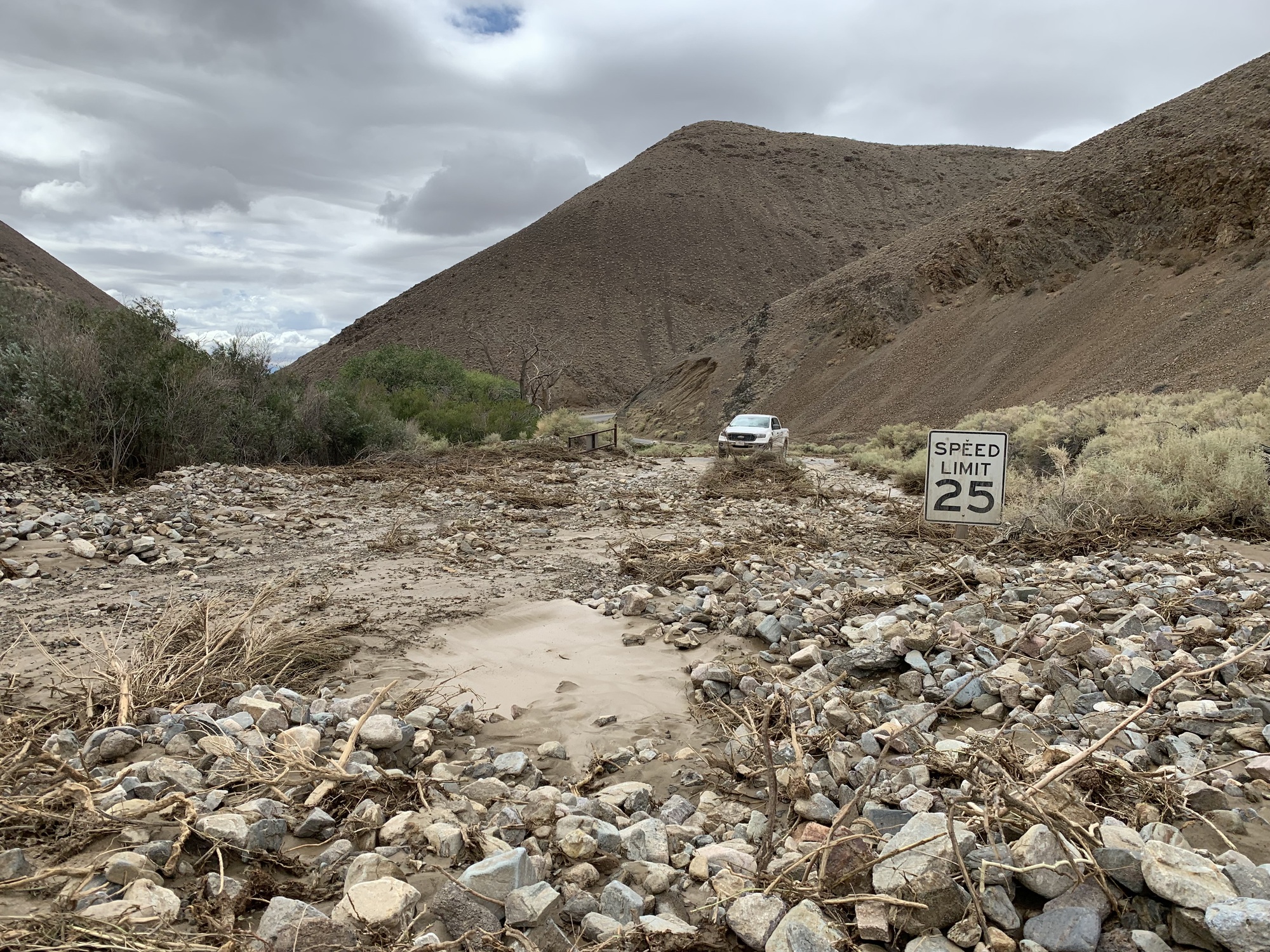 Rocks and mud completely cover a paved road with a "25 Speed Limit" sign just visible above the debris, a white truck in the distance parked at the beginning of the debris and rocky desert hills rising in the background.