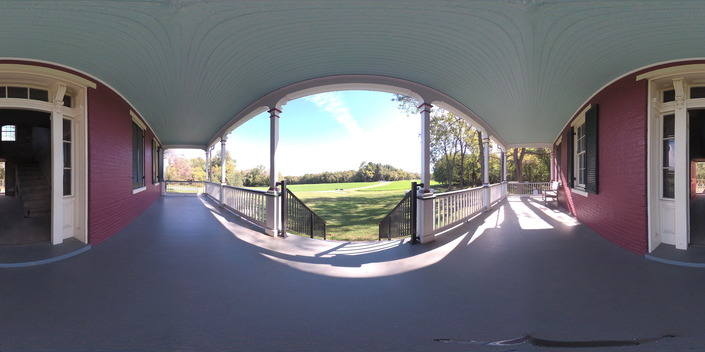 The porch overlooks a grassy field with woods in the distance; a railing surrounds the porch, and stairs lead down to the grass. Behind is the entryway to the house. 