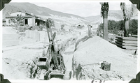 This is an historic black and white photograph from the Scotty's Castle Historic Photograph Collection, Death Valley National Park of power shovel in trench. Tunnel known as Multipurpose Facility (MPF) Tunnel runs from  Scotty's Castle Main House to Hacienda. Looking east towards Stables. Possibly Albert M. Johnson and Bessie Morris Penniman Johnson standing in front of Hacienda. May 1931. Photographed by Mat Roy Thompson.