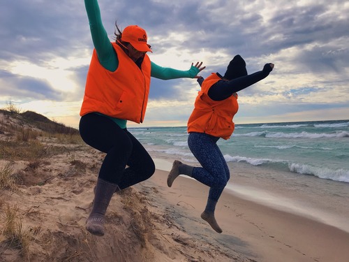 Two women in bright orange vests jump off the edge of a sand dune