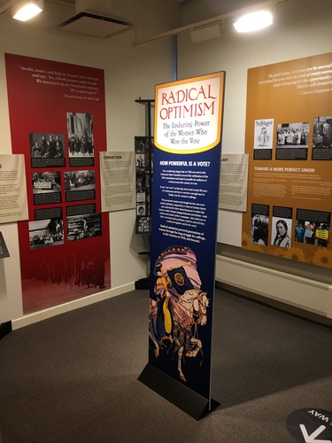 Red and yellow exhibit panels and a free standing banner in the center of the floor with a drawing of a person in yellow, white, and purple holding a long, narrow heralding trumpet with a purple banner hanging off it that reads "Votes for Women" who is riding a white horse. 