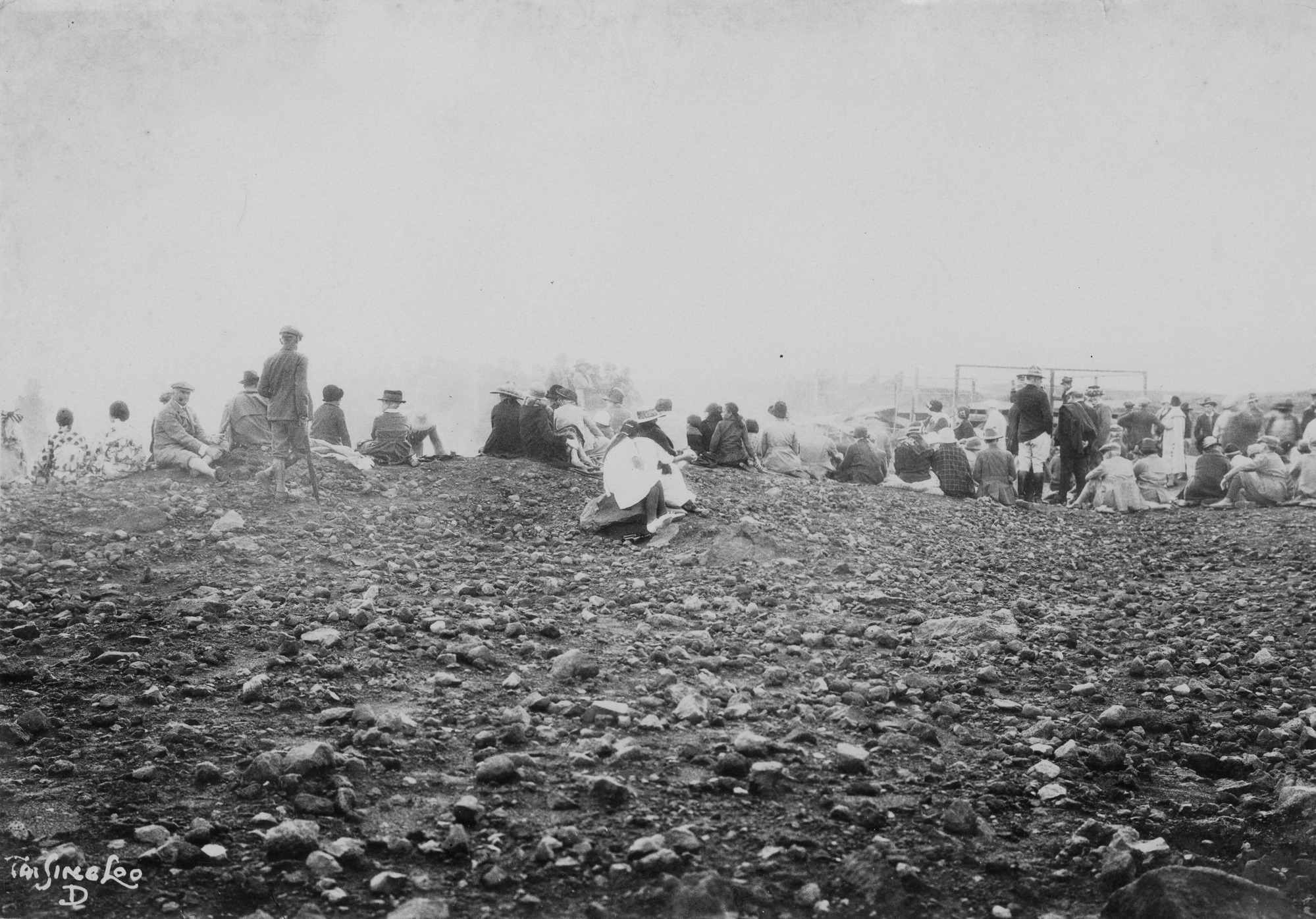 Black and white photograph of approximately fifty people, mostly sitting, looking away from the camera into a large cloud of steam. The crowd is formed in a loose horizontal line across the middle of the photograph. The bottom half of the photo is rocky ground. The top half of the image is almost entirely steam. “Tai Sing Loo, D” is written in the bottom left corner.