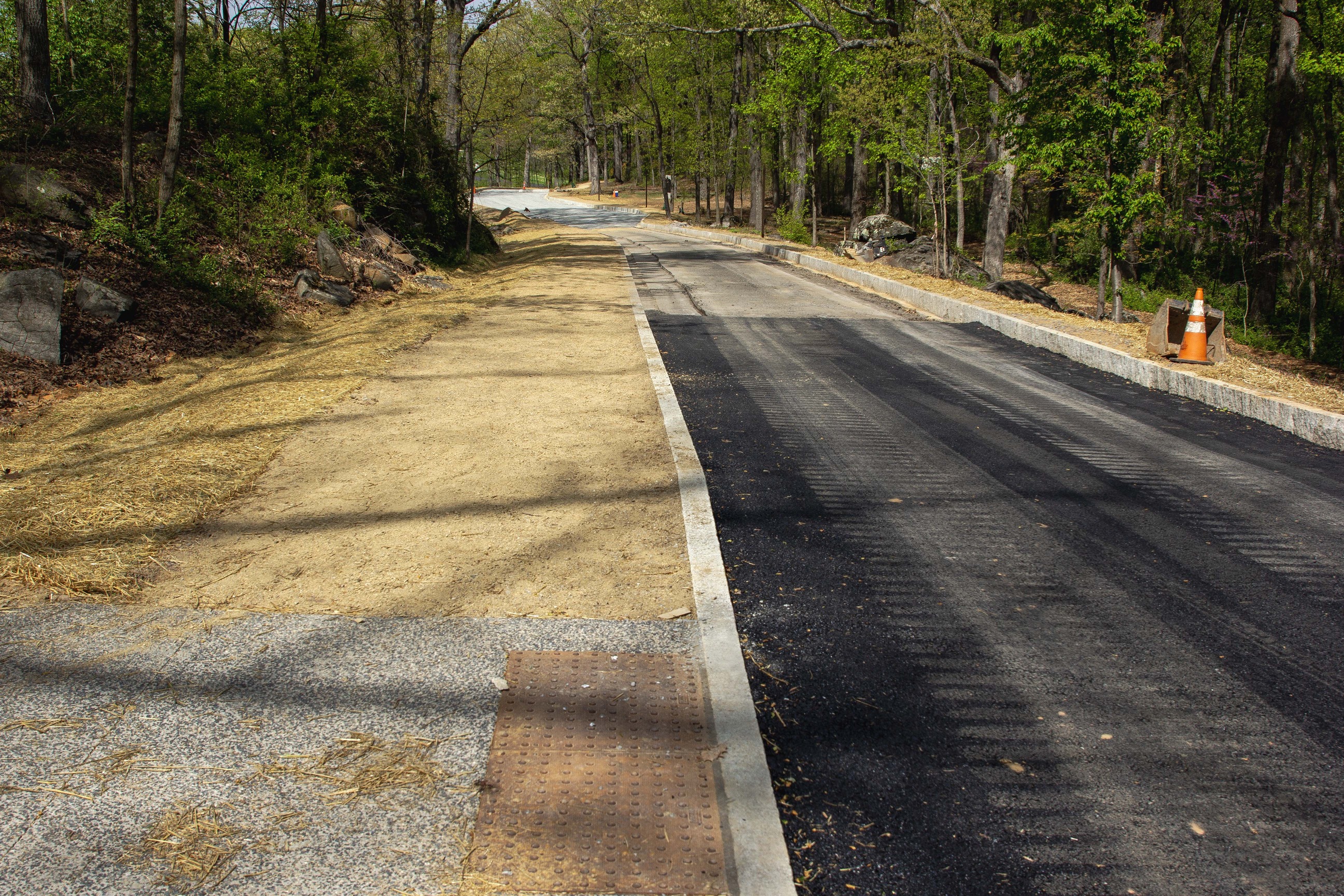 A dirt trail leads along a roadway with recently laid asphalt. Trees and boulders line the sides of the roadway. 