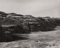 Black and white photograph of a grassy canyon with steep sides made of rounded hills with buildings at the top