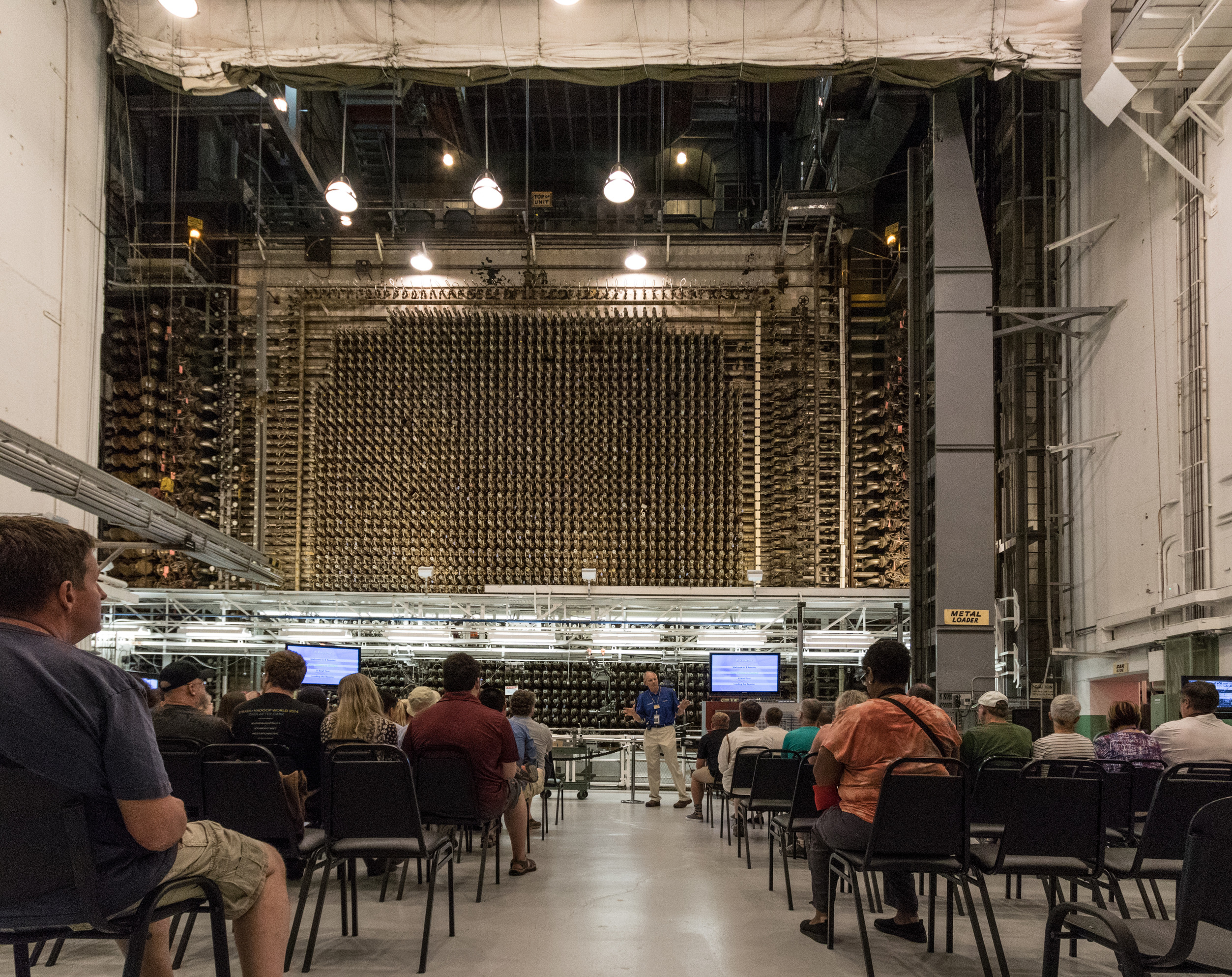 Visitors to the B Reactor sit in rows of chairs in a large auditorium-like room towards the face of the reactor, an enormous wall of hundreds of pipe ends each connected to tubes. A docent presents about the history and science of the B Reactor.