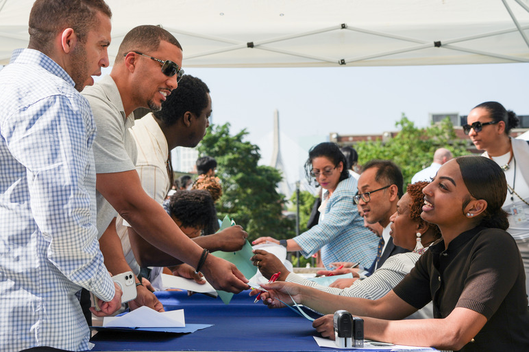 View looking down a table. On the right side of the table, people are seated and speaking to individuals standing across from them on the other side of the table. 