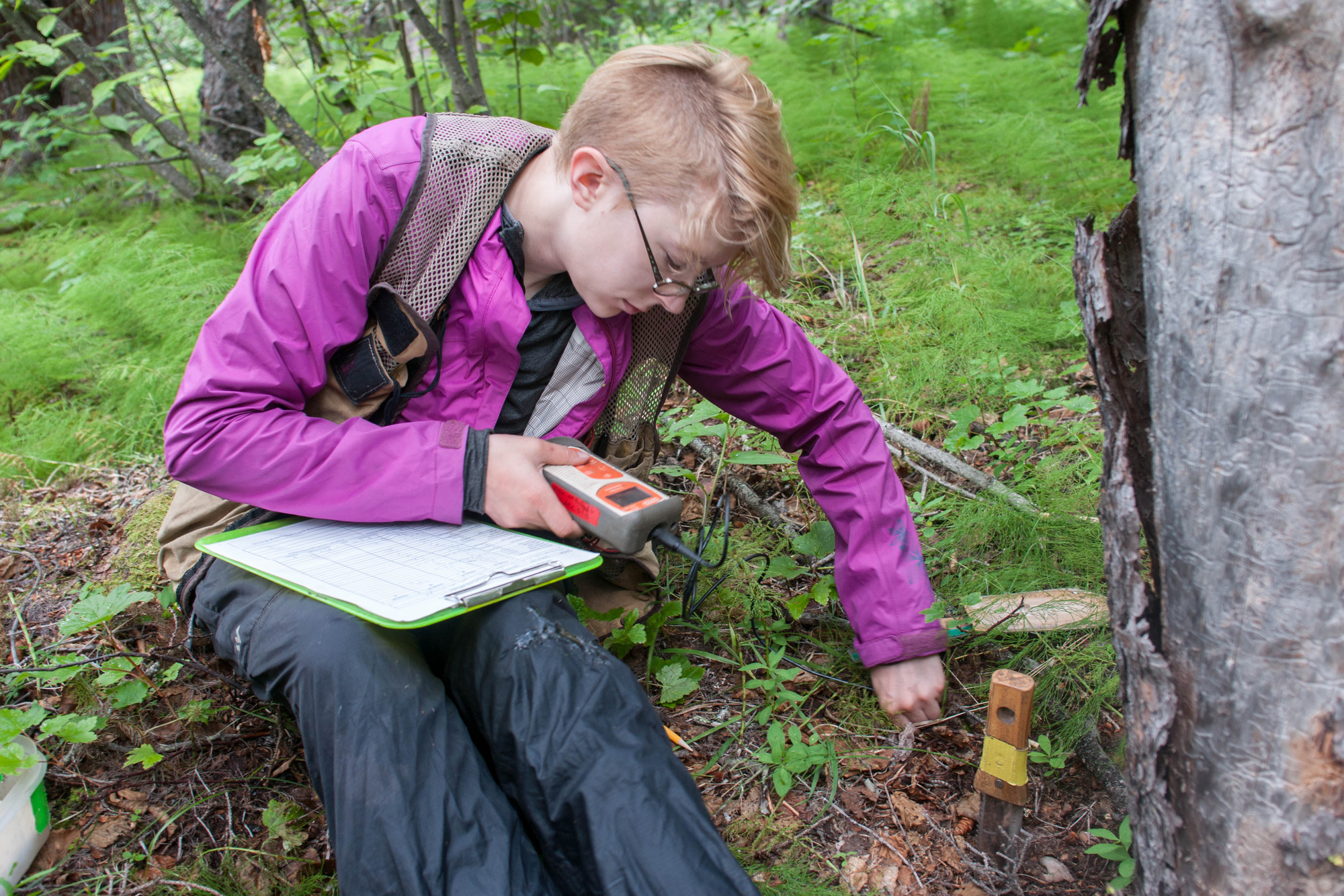 a woman sitting in a forest holding some kind of scientific equipment and a clipboard