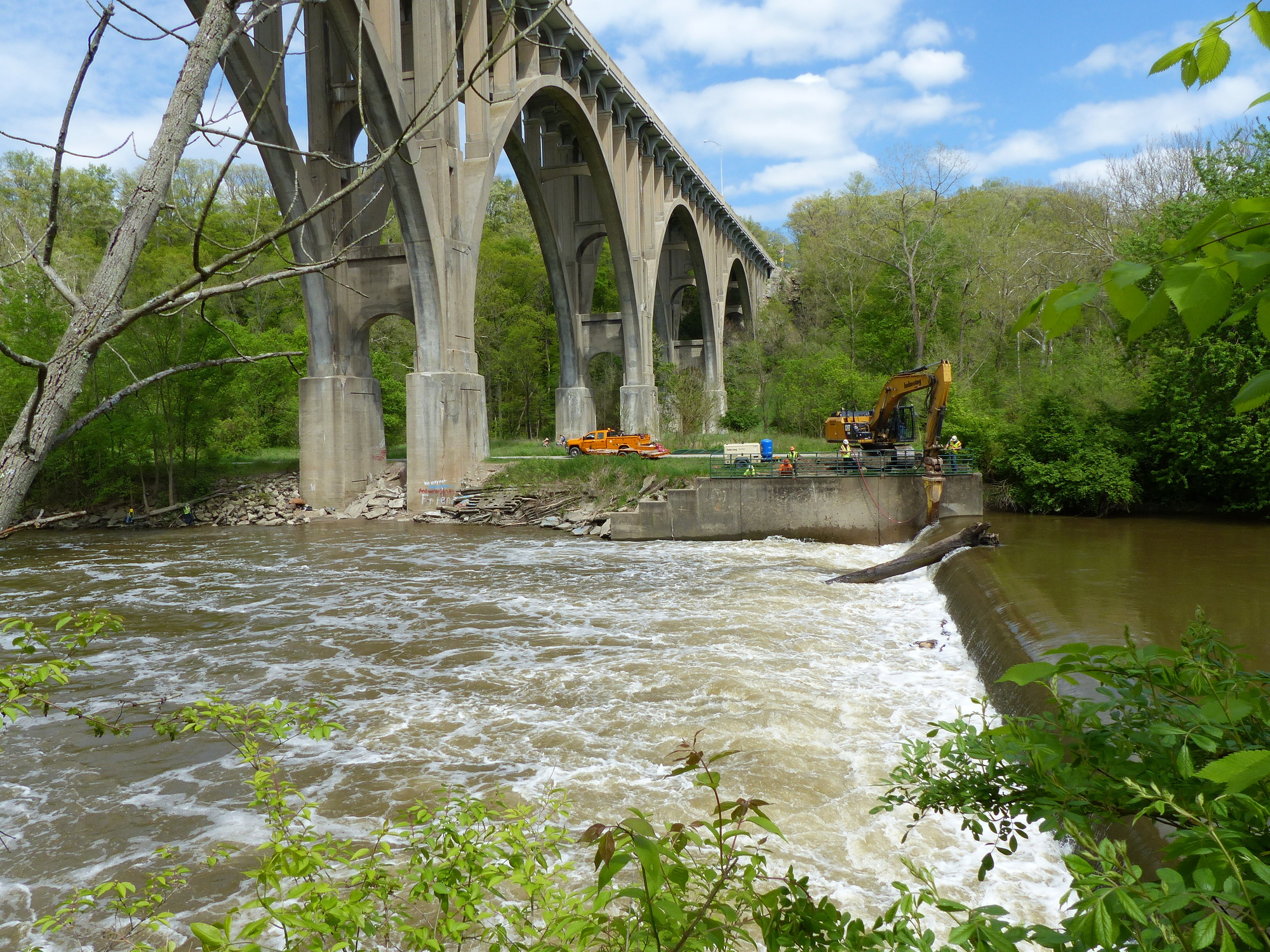 A view looking east of the excavator notching the Brecksville Diversion Dam. The massive concrete arches of the Brecksville-Northfield bridge rise on the left. The calm pool above the dam contrasts with the whitewater below.