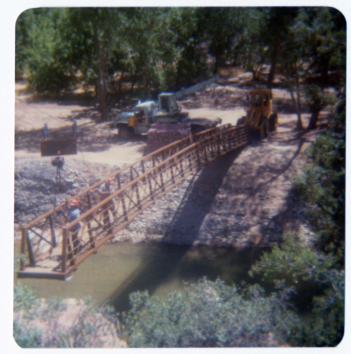 Men on new Grotto footbridge as it is lowered into place by pulley system, crane and backhoe anchoring one end of bridge. Note riverbank revetments.