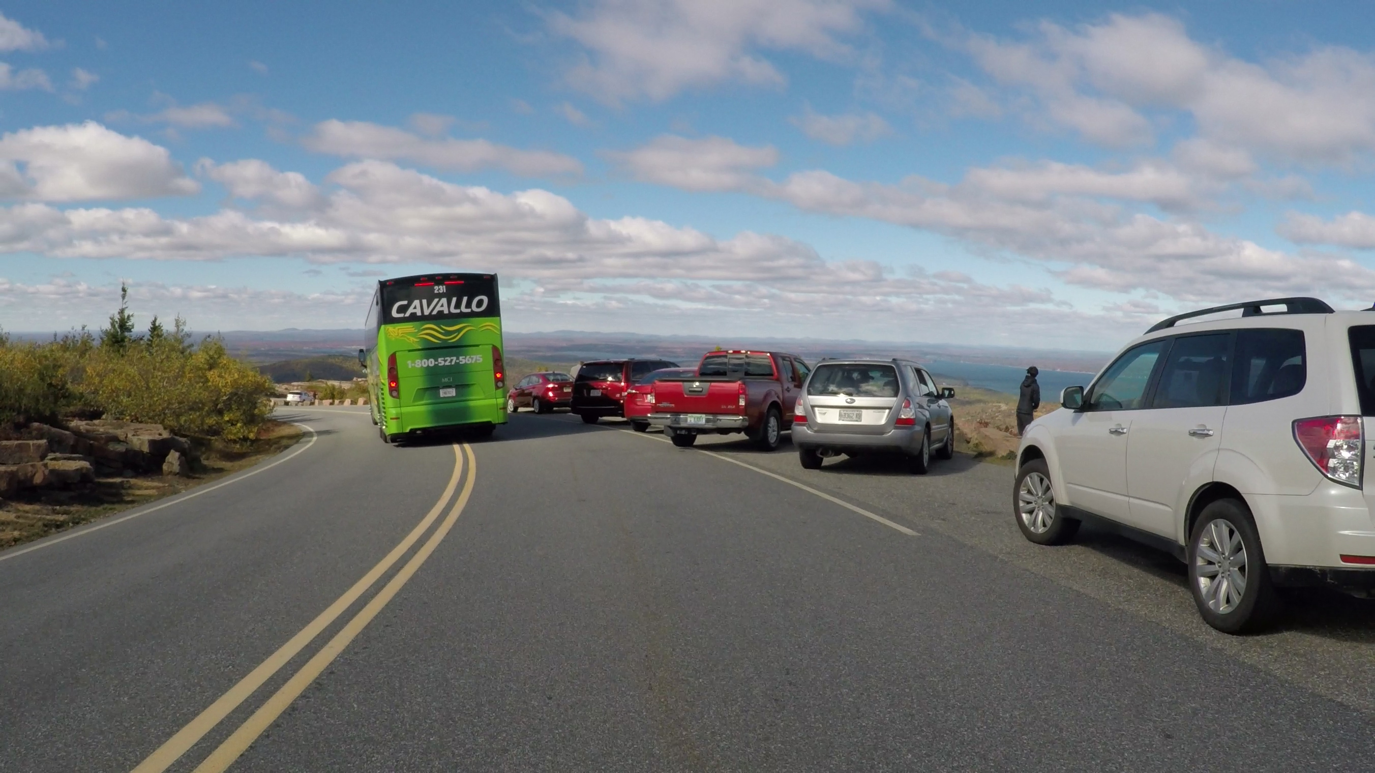 in the center of the image a bus crosses the center lane, on the right side of the image cars are parked in a roadside pullout