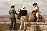 Uniformed woman on a horse speaking with two people at an overlook under a cloudy sky with red badlands in the background.