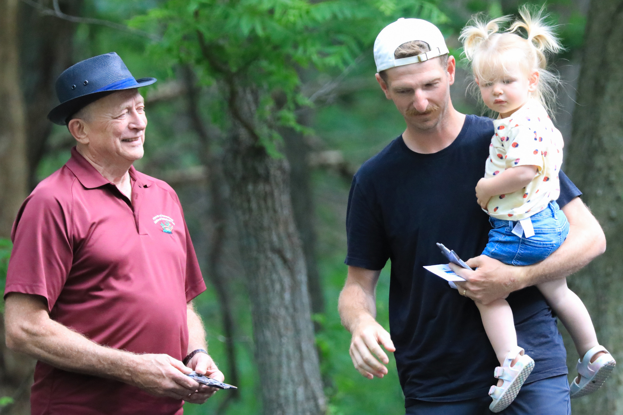Man hands a small trophy to young man holding a little girl in his arms.