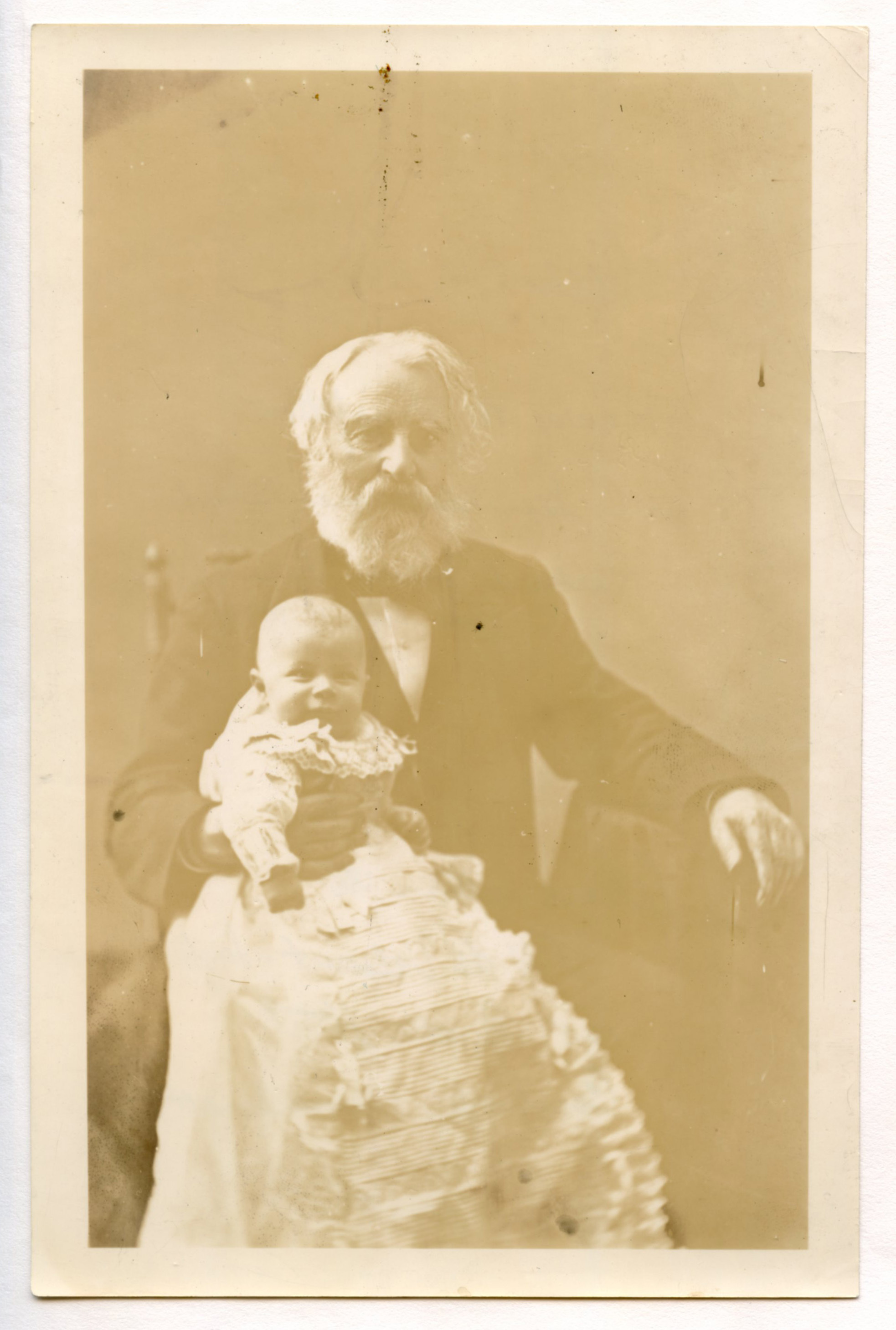 Henry Wadsworth Longfellow is seated, holding a very young baby in his lap. Baby smiles broadly while wearing a long white gown. Henry Wadsworth Longfellow's left arm rests on a table. He wears a dark suit and has ear-length white hair and a full white beard.