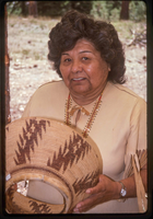 A woman in a Native American attire holds a woven basket. 