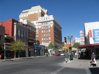 A sunny street scene with historical buildings, shops, pedestrians, and traffic lights in an urban downtown area.