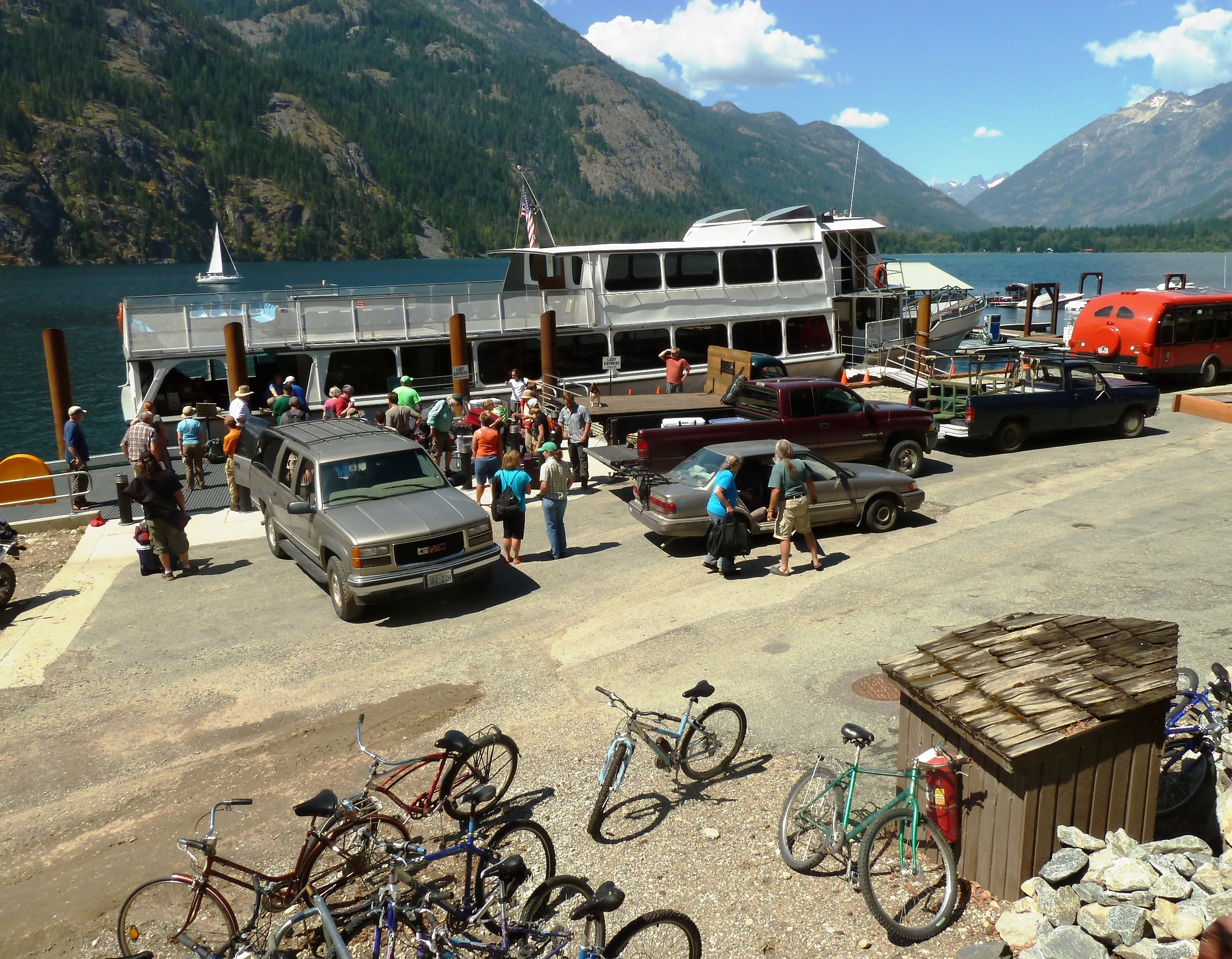 A large silver passenger ferry unloads people at a landing with cars and bikes parked