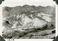 This is an historic black and white photograph from the Scotty's Castle Historic Photograph Collection, Death Valley National Park of a large barn along the length of a wash at base of barren, steep desert mountains. Riparian area of trees and water runs in foreground. Plane prop on right edge.