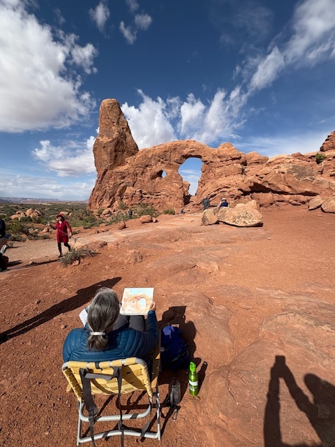 A artist sits in a folding chair painting an arch. Turret arch is visible in the distance.
