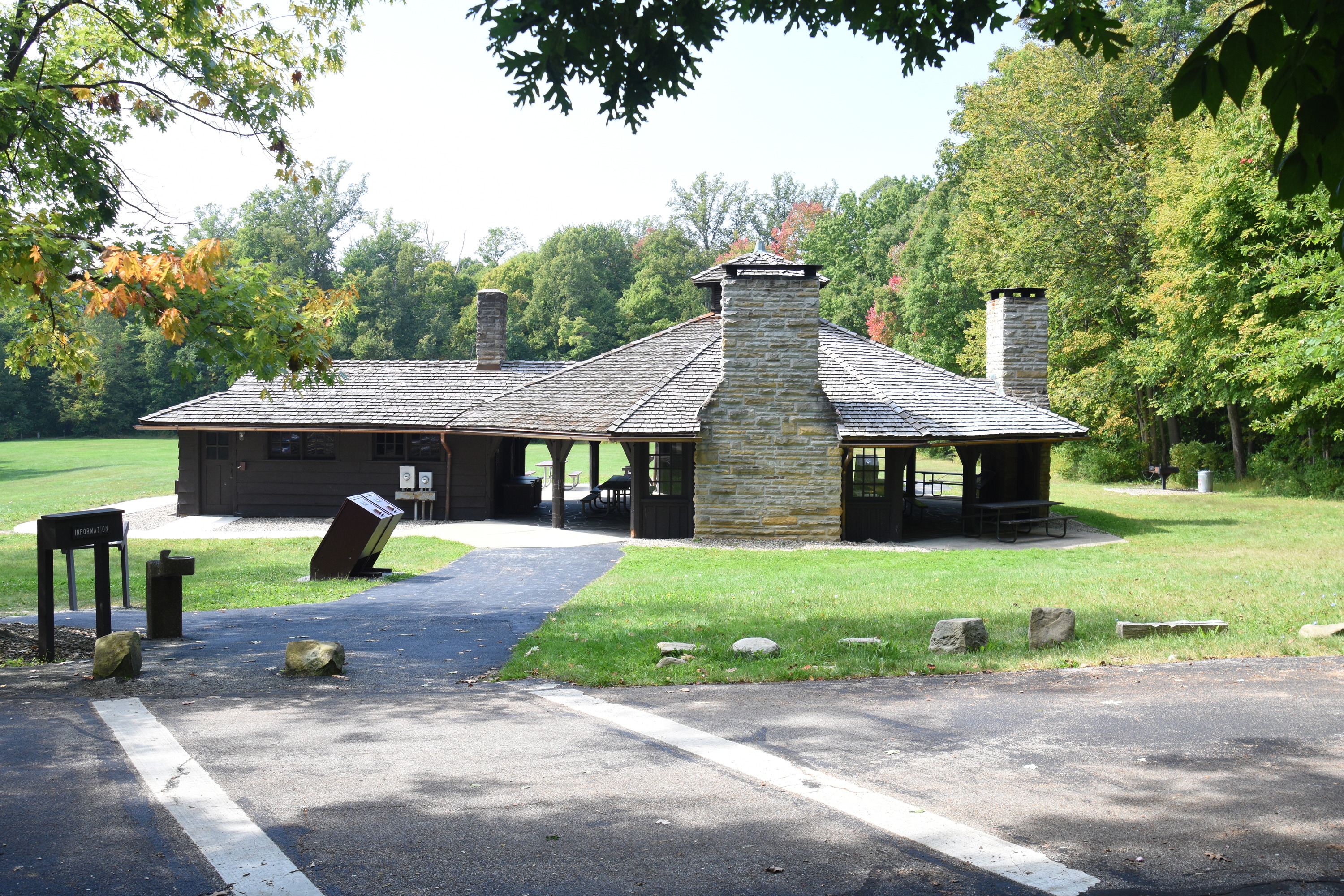 A brown wooden building with two tall, stone chimneys and open walls with brown columns; in the foreground, a crosswalk leads across a paved road toward the shelter; a green field surrounds the building with trees along its edges.