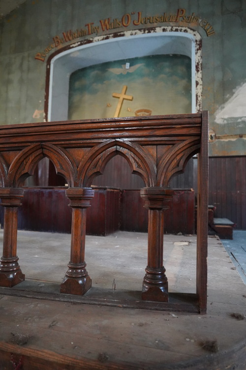 Interior detail of gothic design on pulpit railing, from close up at floor level. Image background shows the dias, alcove, and the back wall mural. Mural depicts a blue sky, clouds, a large gold cross, gold crown and the words "Peace be Within Thy Walls O" Jerusalem Psalm C.xxii"