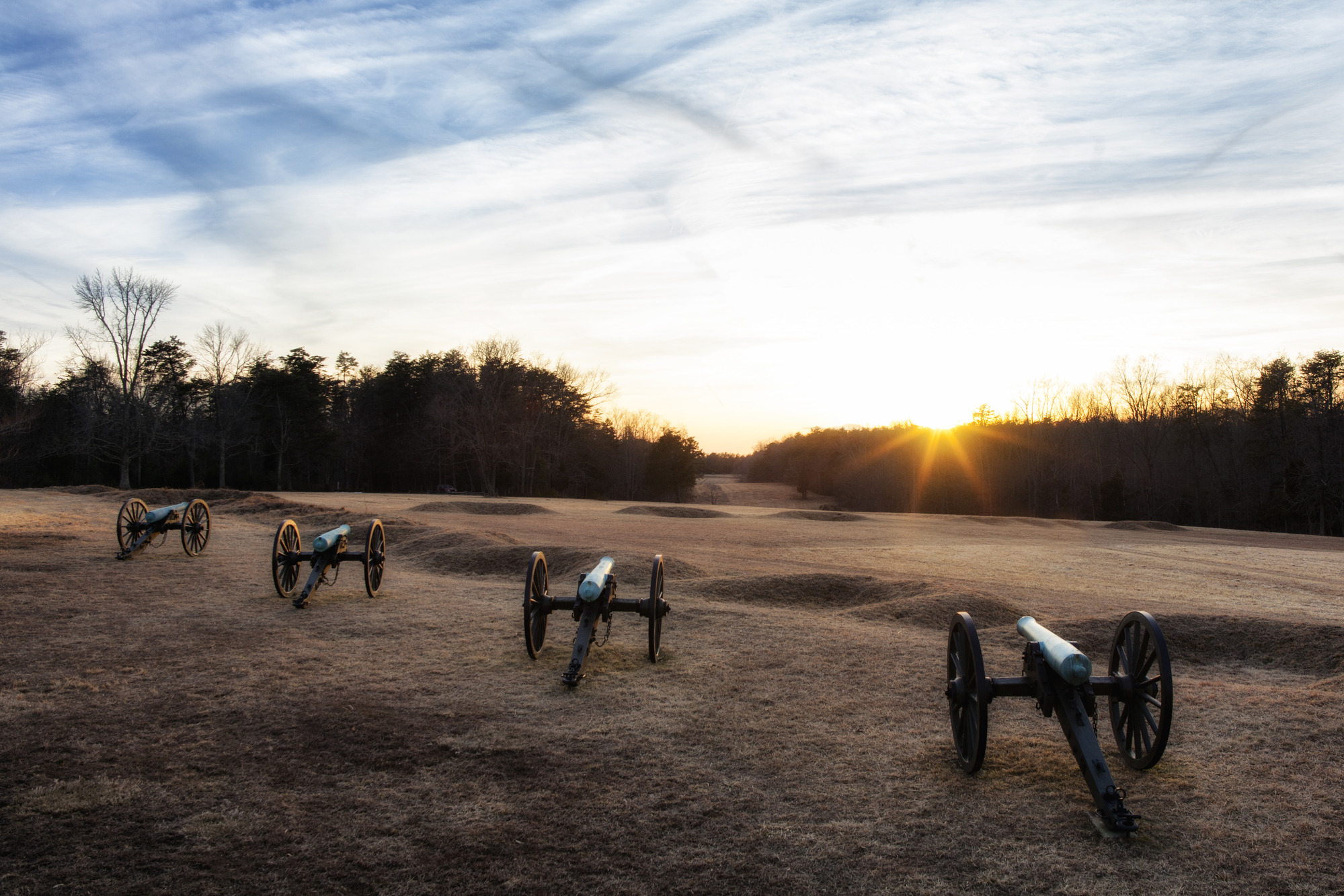 A line of cannons in an open field with earthen mounds, in the shape of half moons, in front of them.