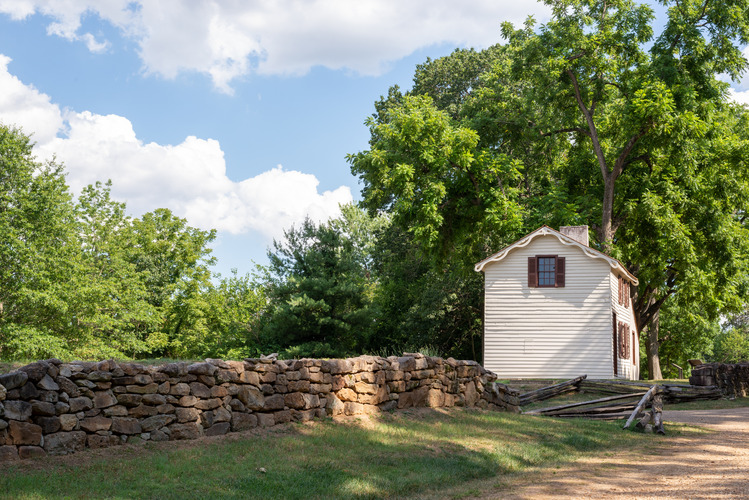 A gravel road lined by a stone wall, leading to a small, white, two story house.