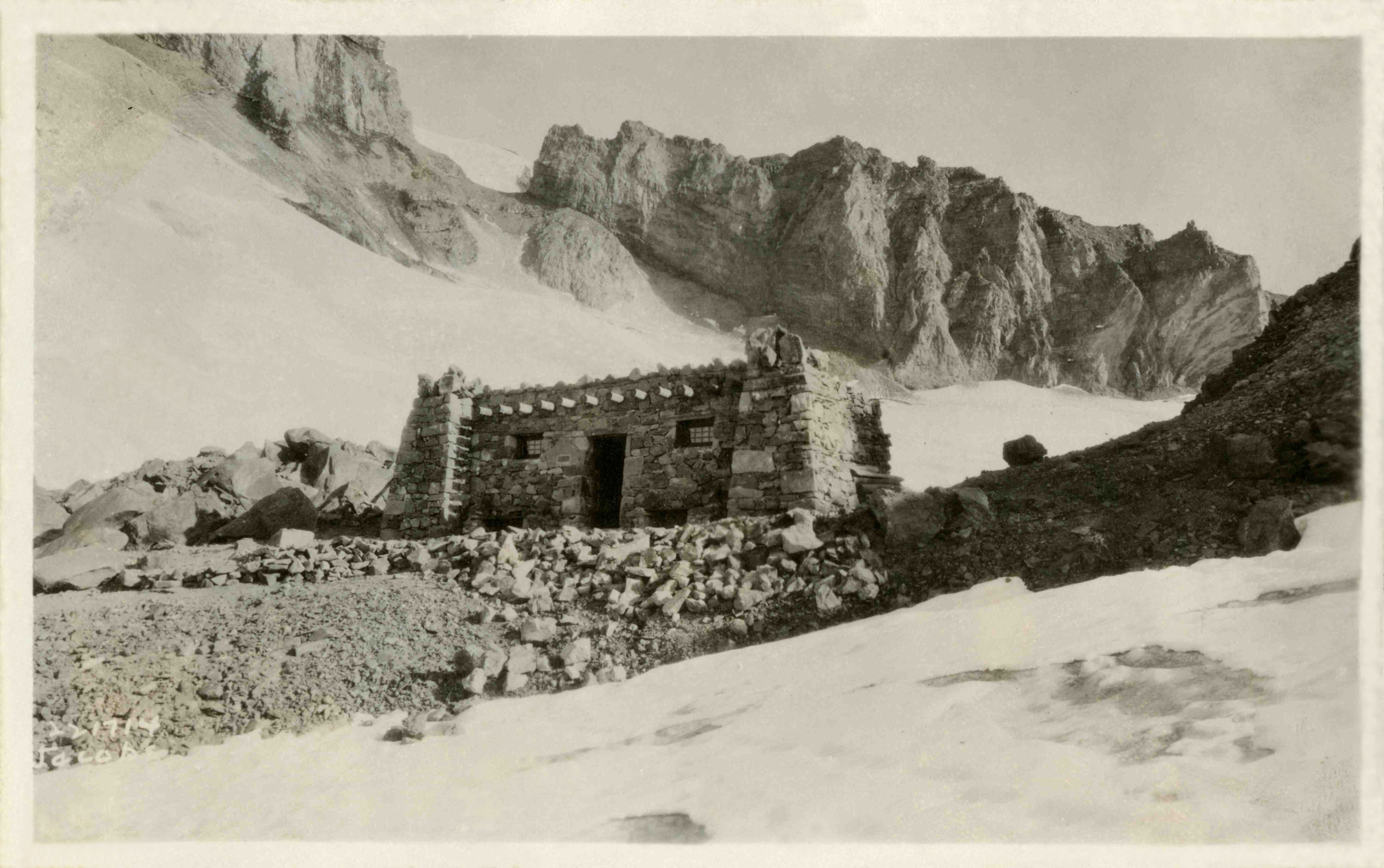 A rectangular stone building with wood eaves on a rocky ridge on a steep glacier-covered slope. 