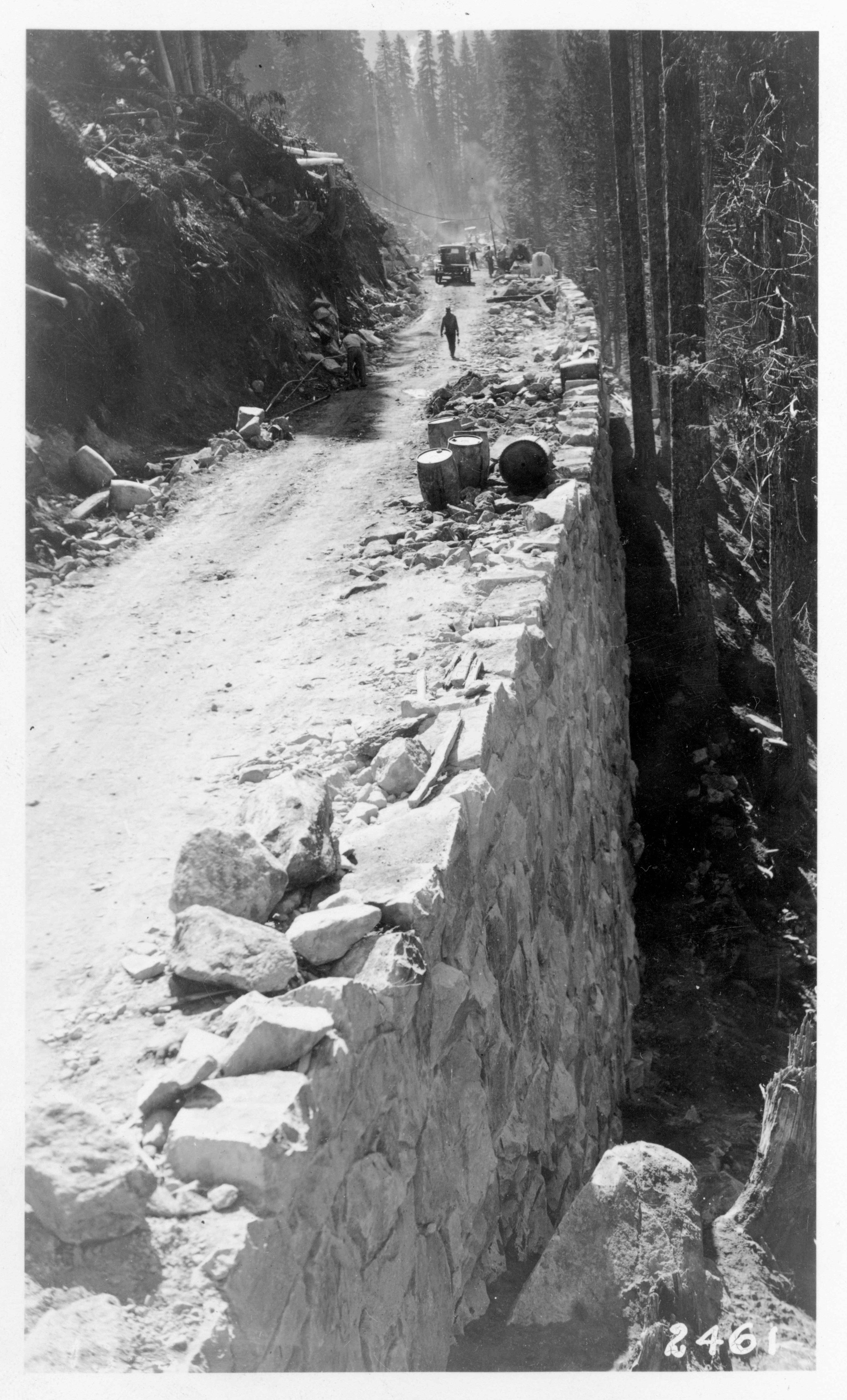 Black and white historic image of a dirt road being cut into a hillside and supported by a rock retaining wall. 