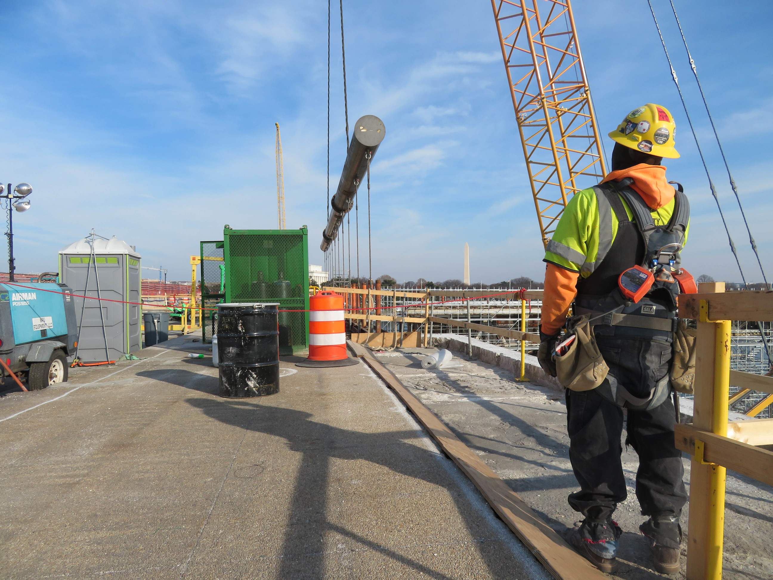 A crane lifts a large rod which is helping lift a large piece of the bridge. A construction work watches