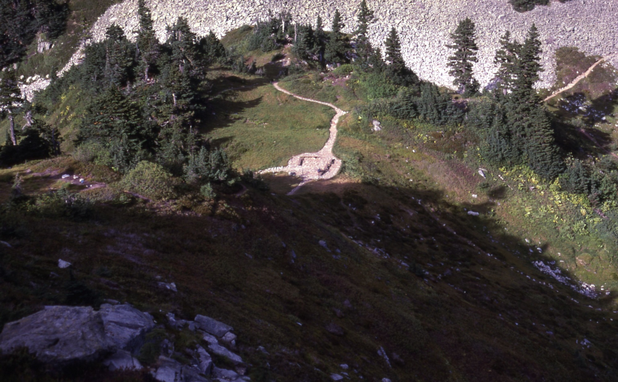 A landscape view down a slope of gentle grassy hills with shrubs, wildflowers, and trees. In the middle is a meadow with a rest area and trails leading away from it. At the bottom is a pit of stones which is cut through by a trail across a rocky mountain slope.