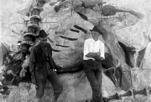 Two men stand in front of an active fossil excavation with dinosaur bones exposed behind them. The man on the left (Earl Douglass) is smiling at the camera. The man on the right in a white shirt (William Holland) has his arms crossed and appears to be looking at him.