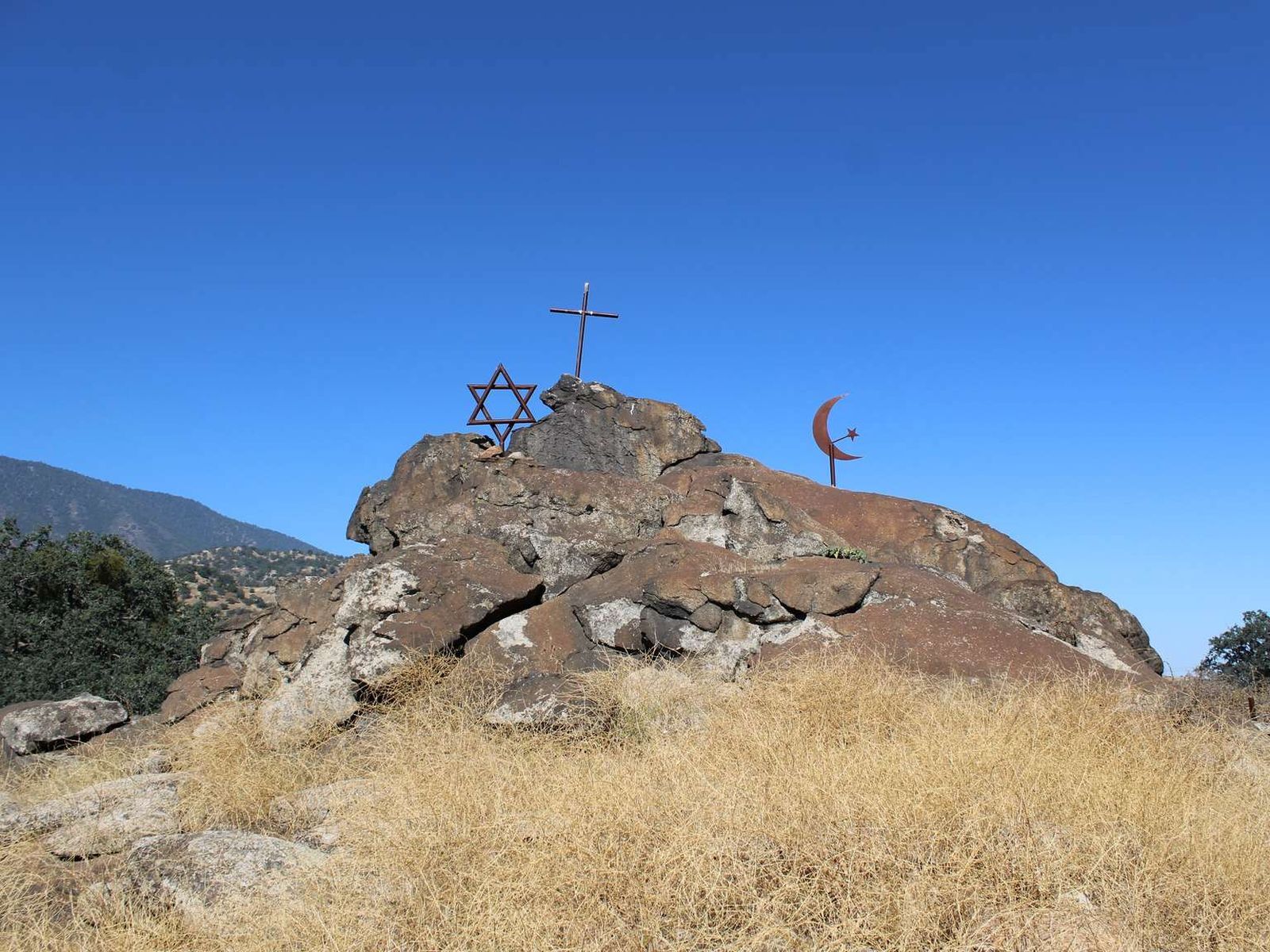 A rock formation with metal symbols of a cross, a star, and a moon. There is dry grass around the rock with trees and hills in the background.