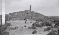 This is an historic black and white photograph from the Scotty's Castle Historic Photograph Collection, Death Valley National Park of woman and child posing in front of mining or milling ruins in desert. Wind blowing woman’s dress. On hillside are remains of stone, terraced walls, wall of a room to the left, some metal milling equipment, and a tall, square tower in center. Extensive foreground of sand, dirt, and desert shrubs.