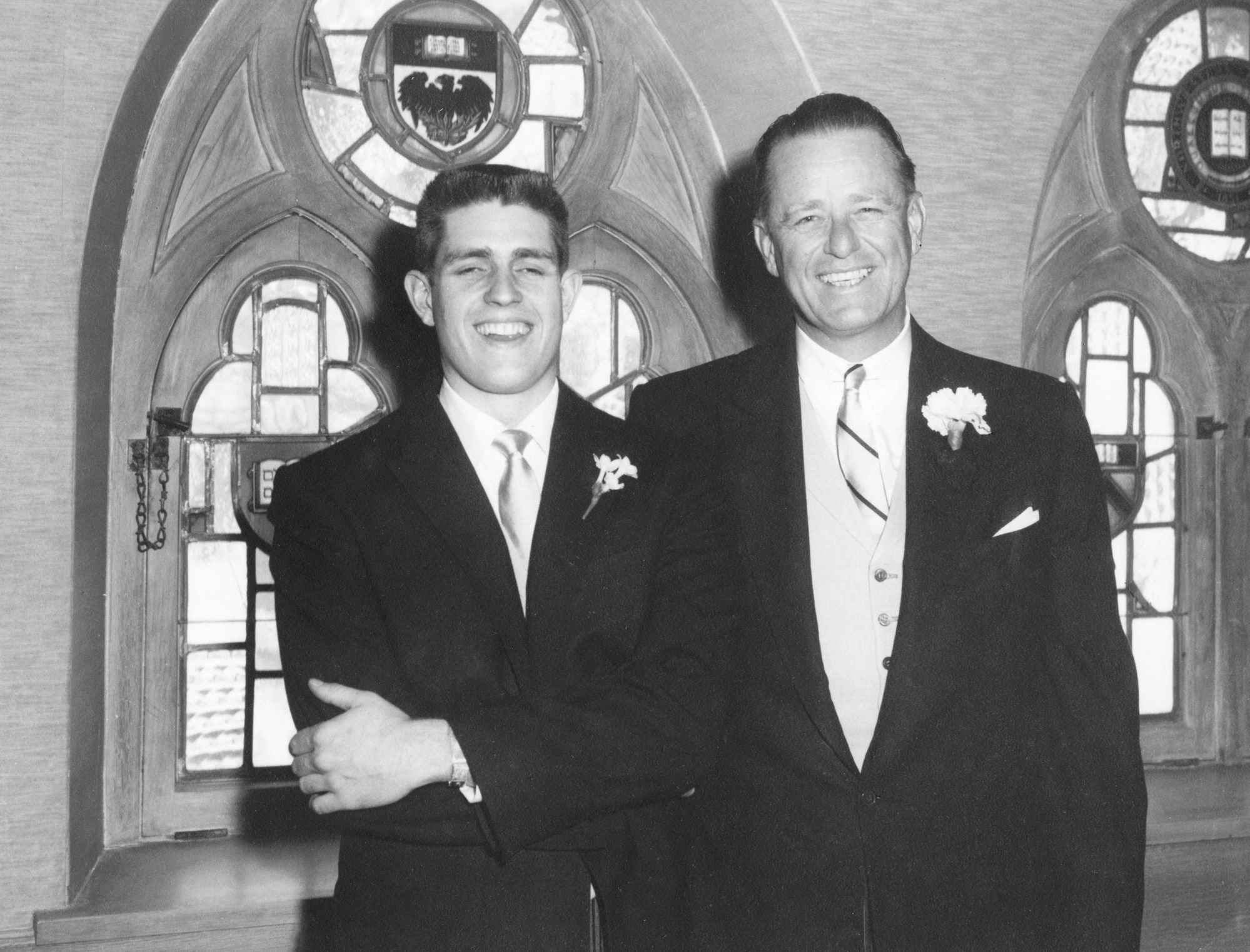 Two men in suits wearing boutonnieres. The man on the left has his arms crossed. 