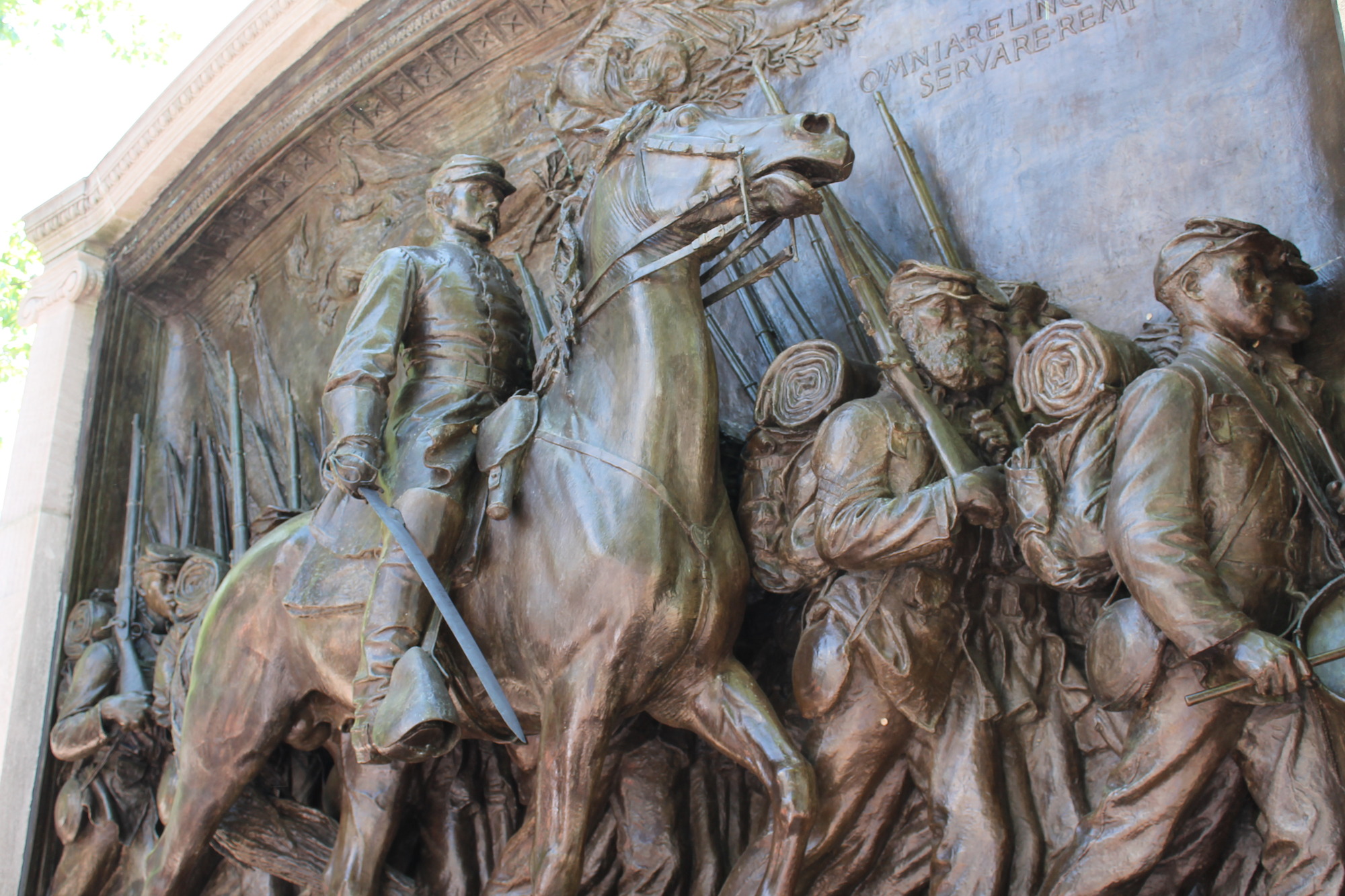 This image shows the statuary portion of the Robert Gould Shaw and Massachusetts 54th Regiment Memorial. There are multiple lines of African American men marching in line with their equipment like muskets, drums, and other items in their hands. On their backs are packs with bedrolls and other items attached to them. Colonel Robert Gould Shaw is depicted riding his horse beside them. The horse's eyes are wide and his mouth is open. Shaw is carrying a sword in his right hand. The sword points forward and down towards the ground. Above all the men is an allegorical figure of an angel either of death or mercy who is floating in the air.