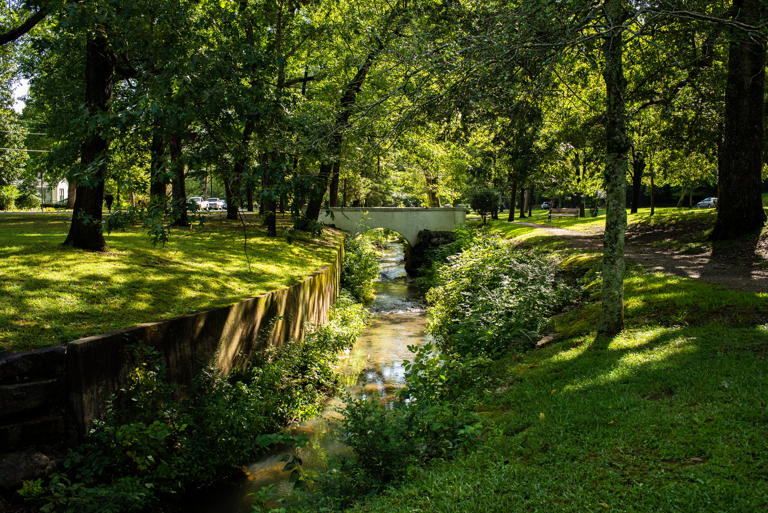 Dappled sunlight shines through the trees and onto the creek. There is a small, white, arched, historic bridge in the background.