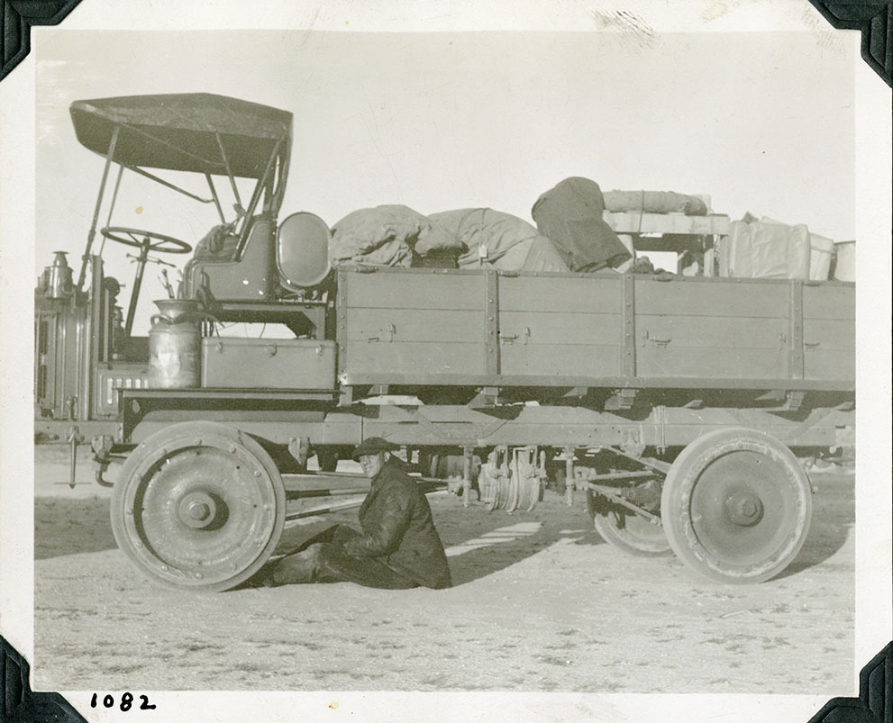 This is an historic black and white photograph from the Scotty's Castle Historic Photograph Collection, Death Valley National Park of a man on ground behind front tire of very large truck. Truck bed filled with supplies, many covered with canvas. Large toolbox and cream can on running board. Open cab with canvas cover. Number in black ink in lower left corner.
