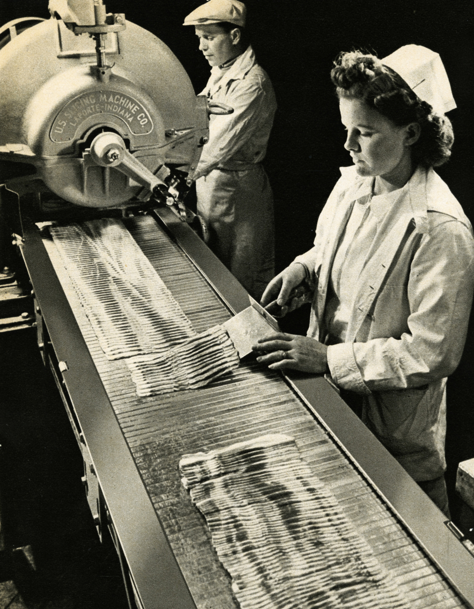 Black and white photograph of a woman slicing bacon on a conveyor belt alongside a man