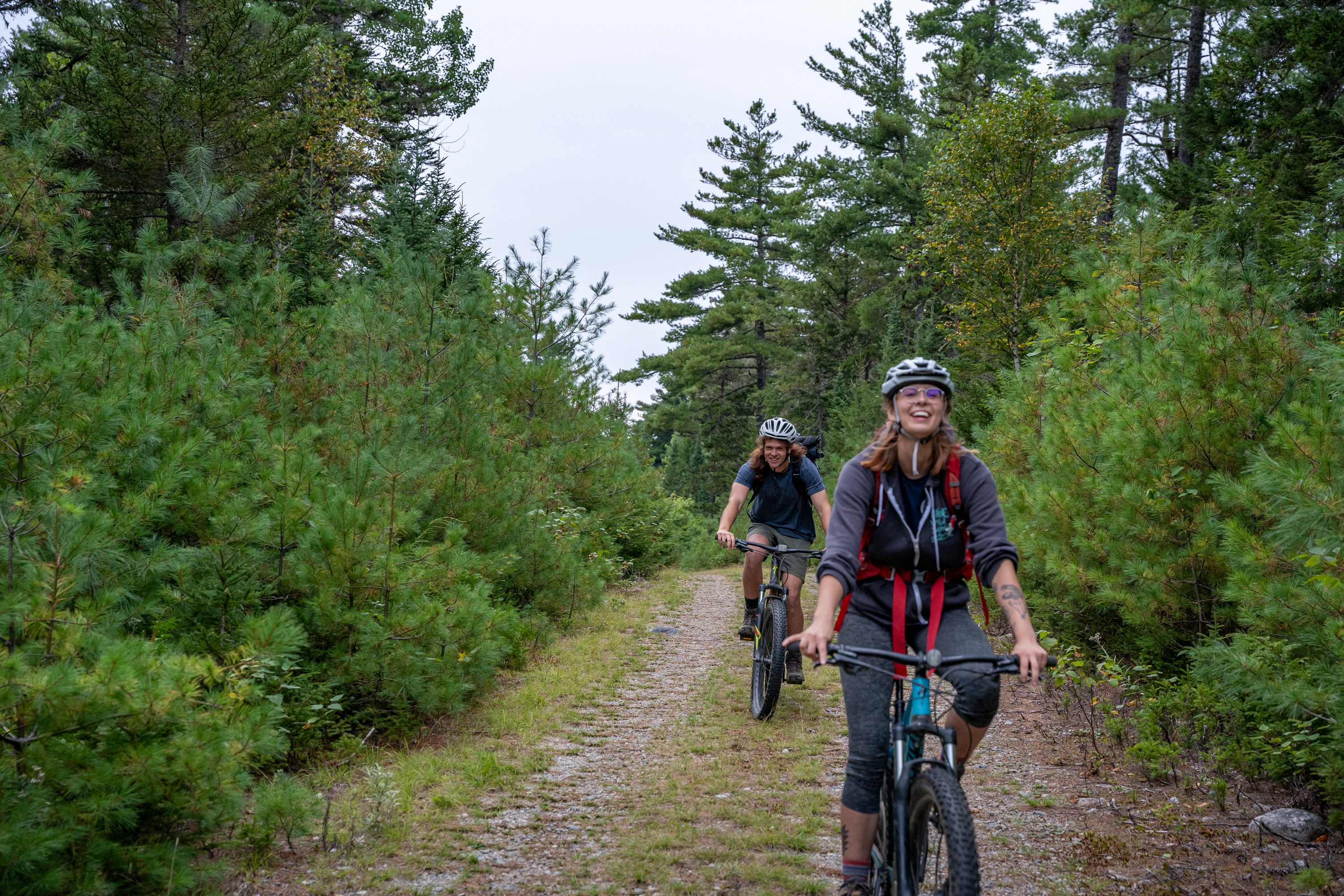 One biker in the front with a white helmet on and dark clothing is followed by a second biker with a white helmet on. They are smiling and riding on a dirt road with lush green trees surrounding them.