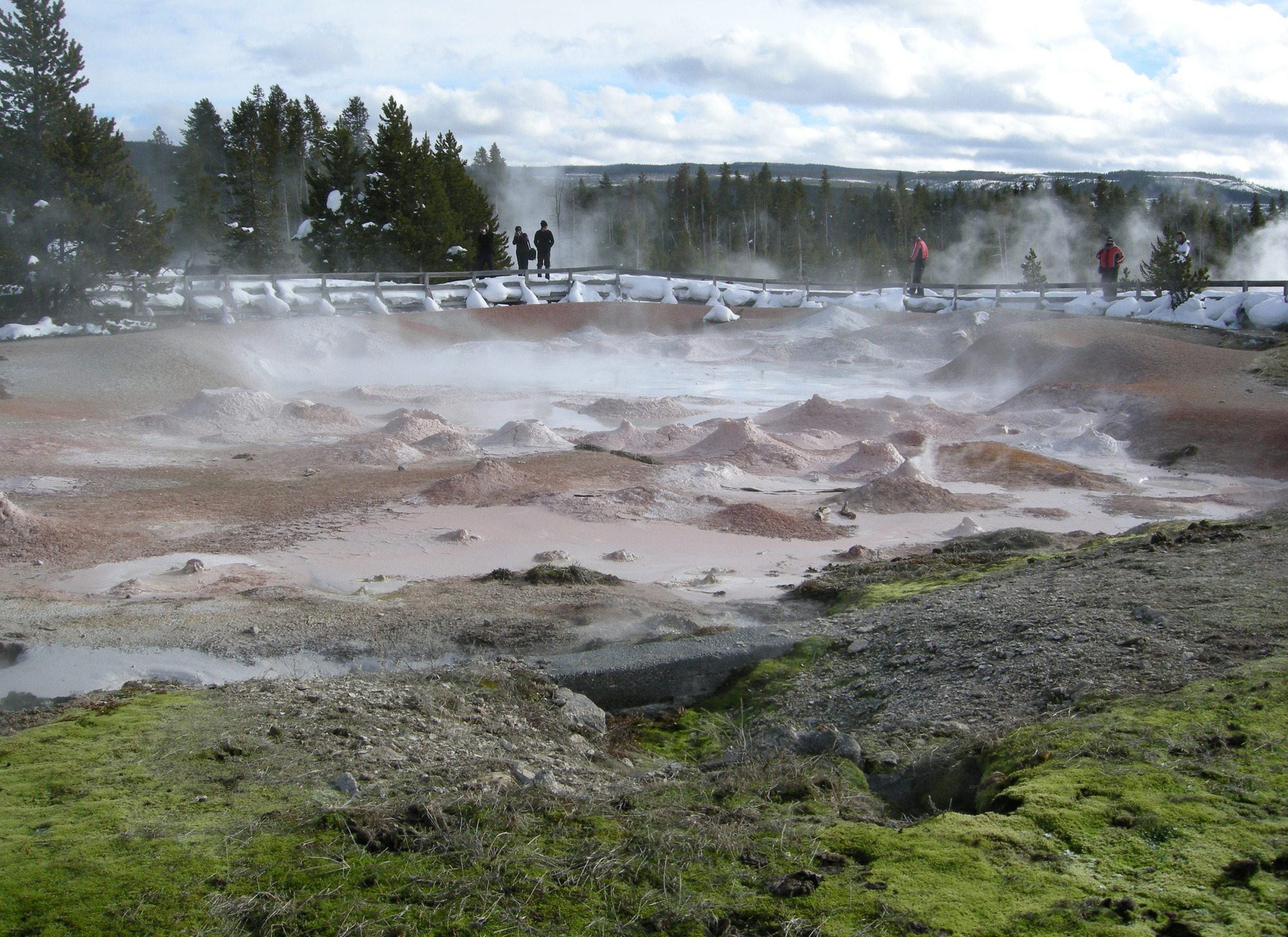 Looking over a bubbly, steamy mudpot at several people standing on a snow covered boardwalk.