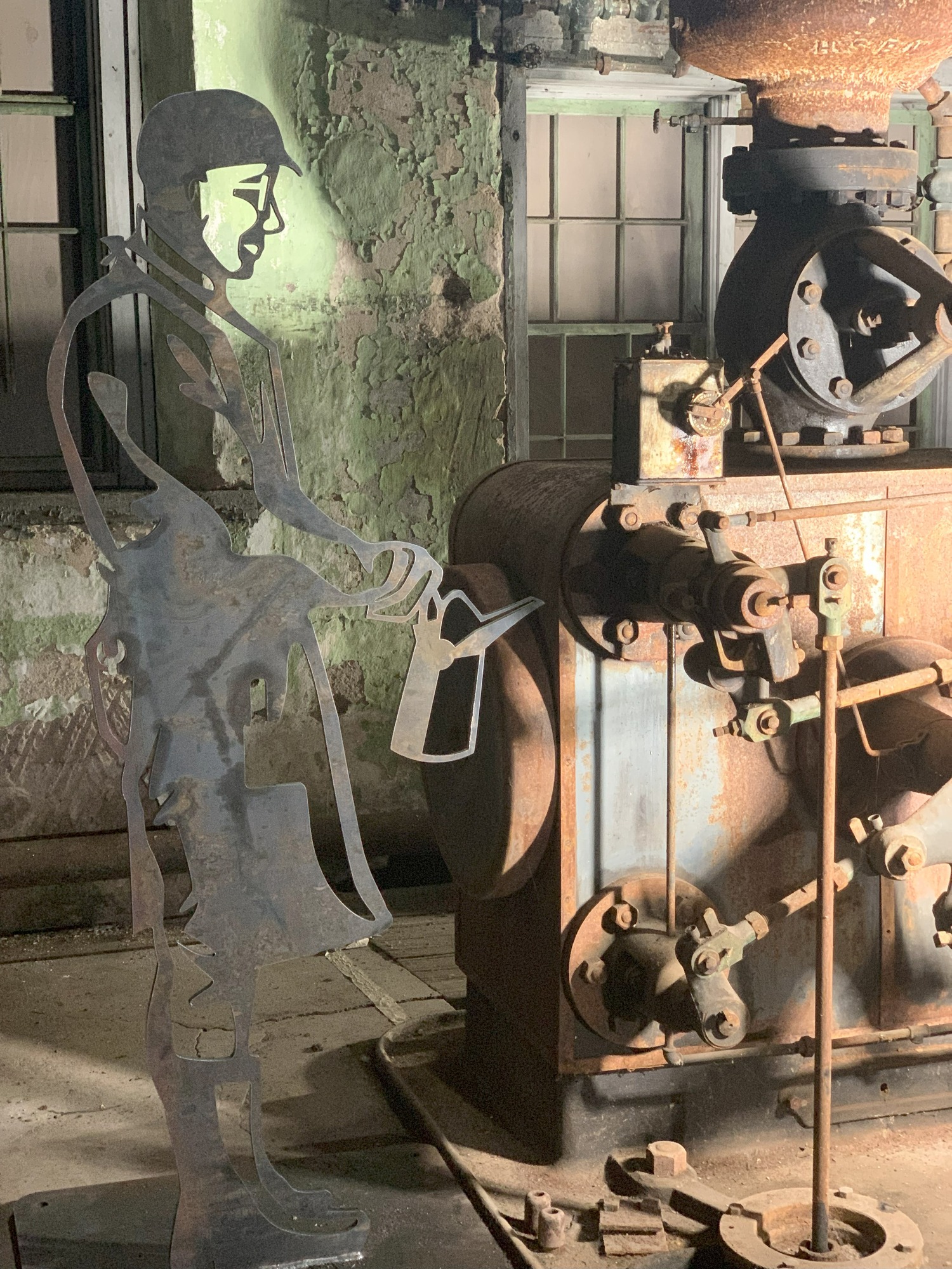A metal sculpture of a man holding an oil can stands near a piece of steam driven equipment.