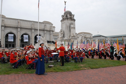 A public event in a grass and brick plaza includes a uniformed band, a vocalist standing at a microphone, and a collection of flags in front of a granite fountain.