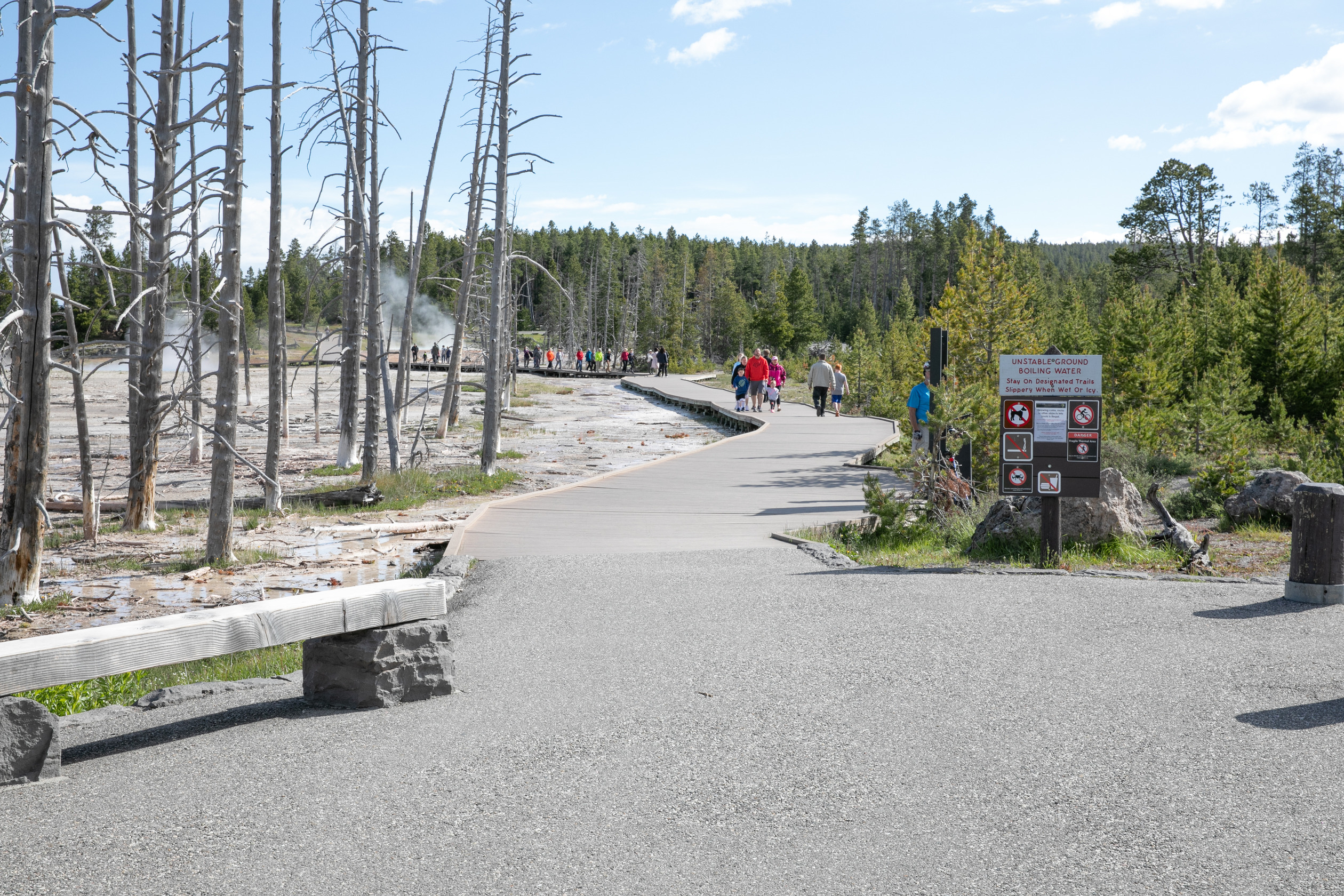 People walk on a boardwalk towards thermal features that are putting steam into the air.