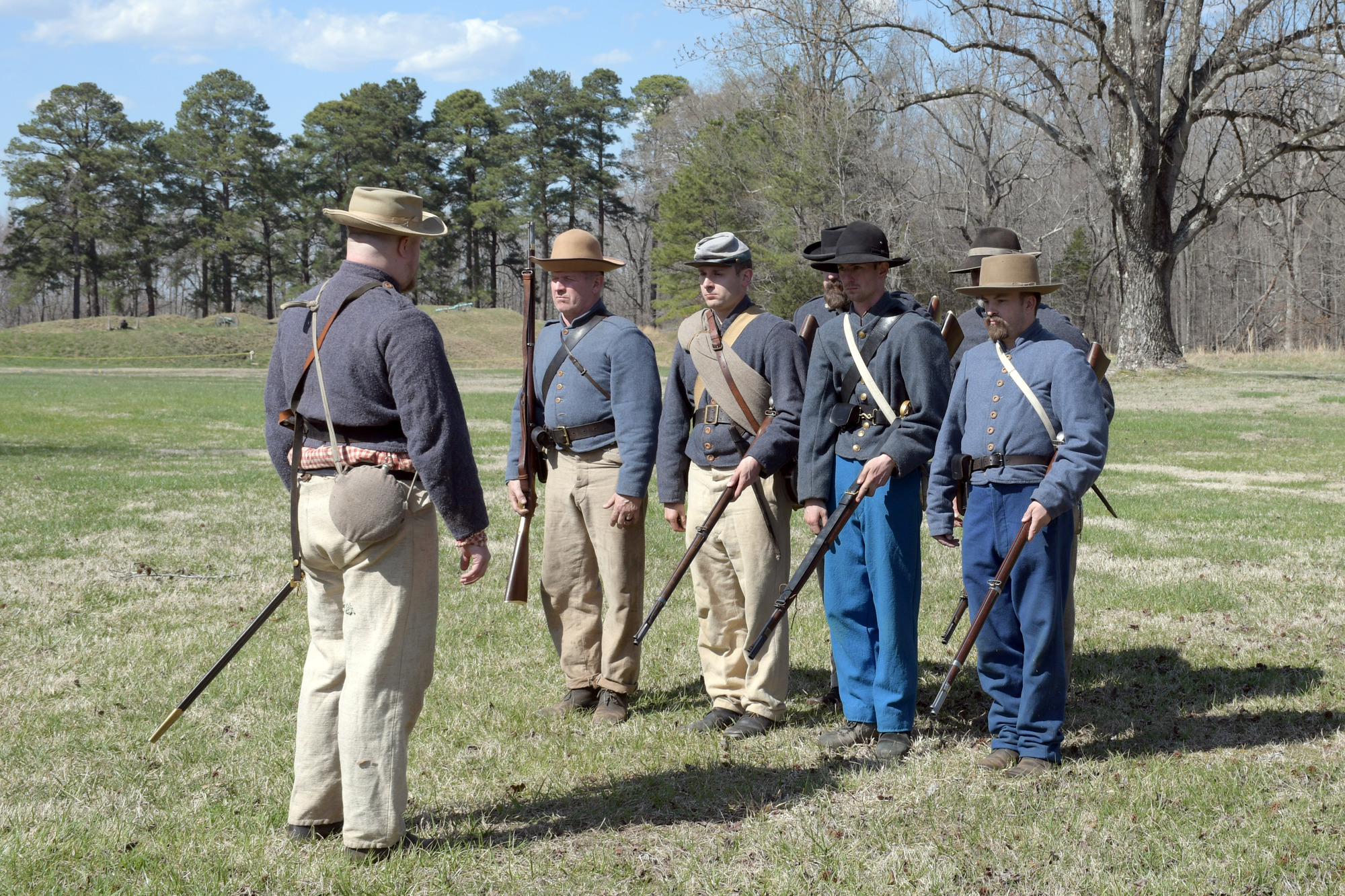 Five men wearing Confederate uniforms stand in formation in front of a 6th man. Four of the men in formation are holding their rifles in their left hands with the barrels pointed toward the ground at a 45-degree angle. The 5th man in formation is standing at attention.