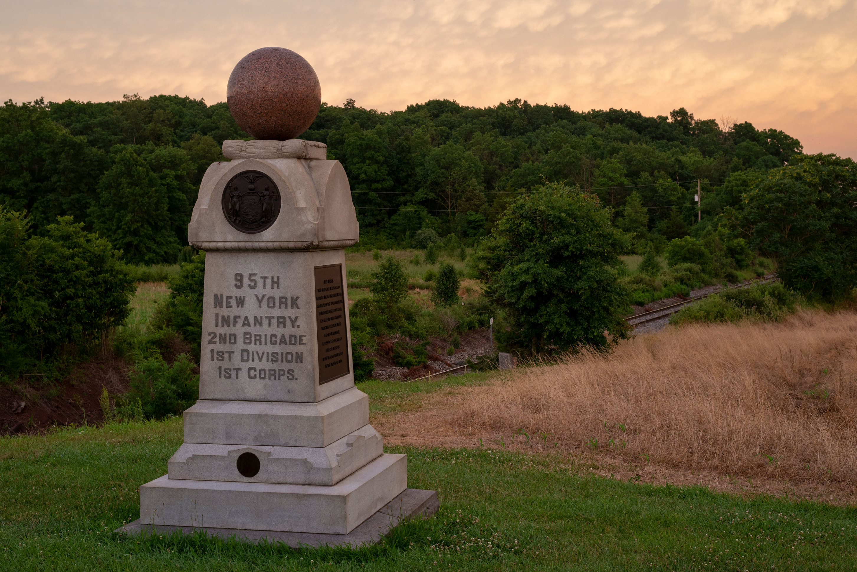 A rectangular monument topped with a sphere perched on a grassy knoll near a railroad track.